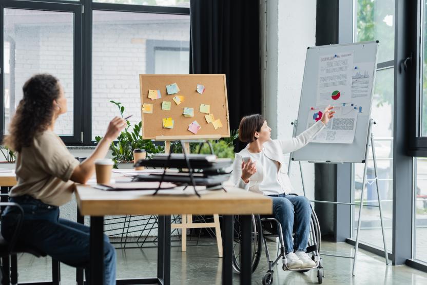 two women in a conference room, one sitting on at a table and another wheelchair user drawing on a whiteboard