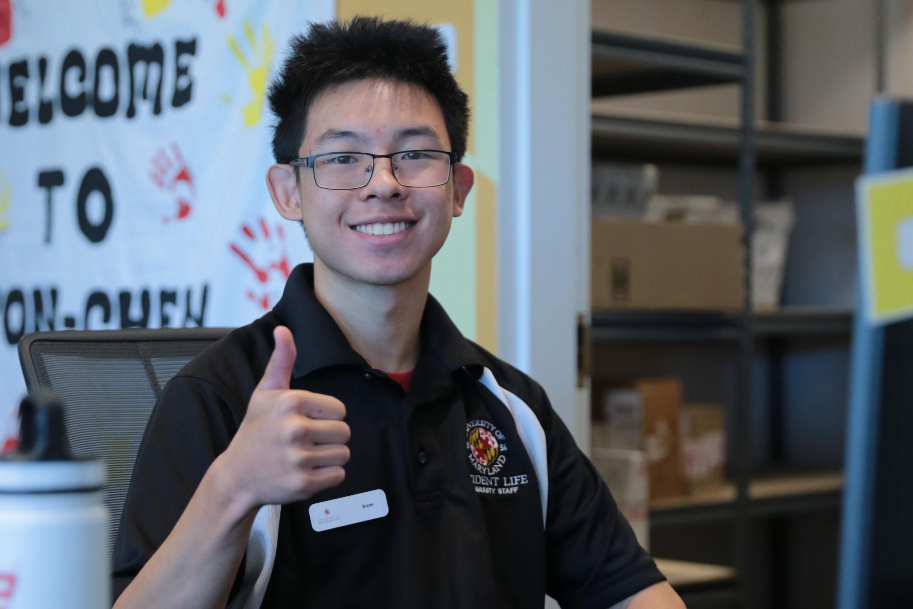 a smiling student employee behind a service desk wearing a black uniform shirt and giving the camera a thumbs up
