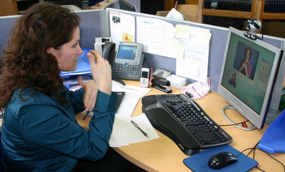 A person at her workplace, communicating with someone via a Sign Language interpreter, shown on-screen, using a webcam and a video call program on her computer. photo credit: Sign Video